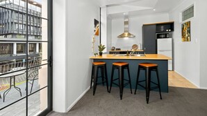 A modern kitchen with a central island, three bar stools, a stainless steel sink, and a view through a glass door to an outdoor area. - Heart of Hamilton Loft – Light, Luxury & Location (Hamilton)