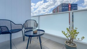 Un balcon avec deux chaises et une table, un pot de fleurs et une vue sur un bâtiment moderne orné d’un logo rouge.