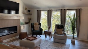 Un salon spacieux doté d’une cheminée, de sièges confortables et de grandes fenêtres offrant une vue sur les arbres.