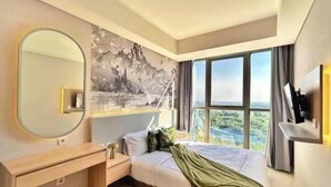 A modern bedroom with a large bed, a bedside table, a mirror, and a view of a mountainous landscape through the window. - TenLira By SanLiving - 1BR - PIK (Jakarta)