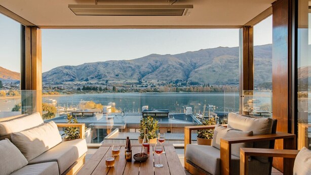 A modern balcony with a view of a lake and mountains, furnished with a sofa, chairs, and a wooden table with drinks.