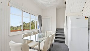 Een kamer met een eettafel met glazen blad, zes stoelen, een witte koelkast, een trap en een raam met uitzicht op groen.