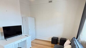 Een moderne woonkamer met een flatscreen televisie op een wit bureau, een zwarte bank met kussens en een hanglamp.