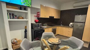 Une cuisine moderne avec des armoires en bois, un comptoir noir et un four encastré. On y trouve une table à manger avec quatre chaises grises et une petite table ronde avec un set de table. Un téléviseur à écran plat est fixé au mur, au-dessus de la cuisine.