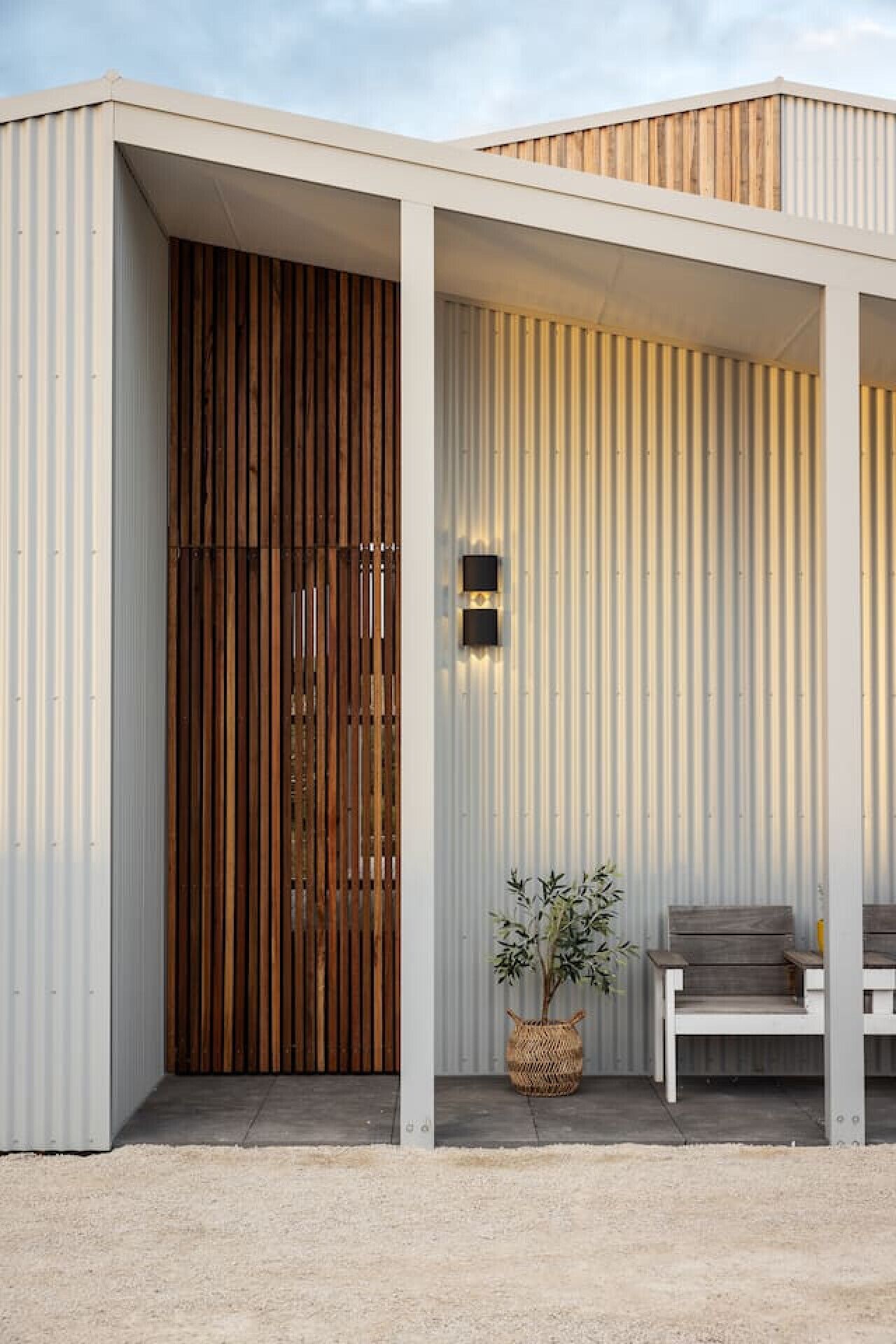 A modern building with a wooden slatted door, a white bench, and a potted plant.