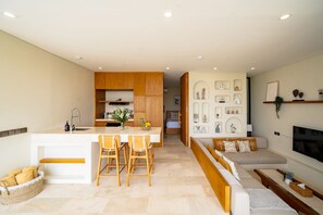 A modern kitchen with wooden cabinets, a central island with bar stools, a dining table, and a living area with a sofa and TV. - Villa Bingin Hills Cinco (Pecatu)