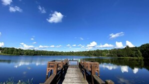 A wooden pier extending into calm water, surrounded by dense greenery and a clear blue sky. - Lakefront Poconos 5BR | Hot Tub • Dock • Near Ski (Blakeslee)