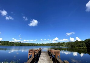 A wooden pier extending into calm water, surrounded by dense greenery and a clear blue sky. - Lakefront Poconos 5BR | Hot Tub • Dock • Near Ski (Blakeslee)