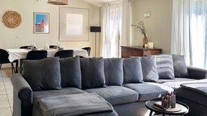 En moderne stue med en hjørnesofa, to hængende fletlamper, et spiseområde med et hvidt bord og udsigt til det fri gennem store vinduer.