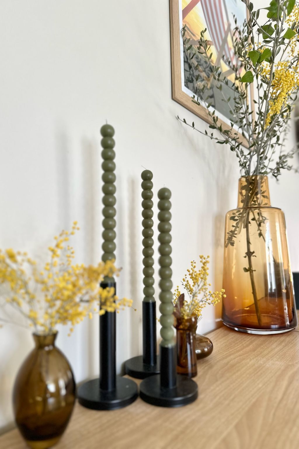 A wooden table with three black stands holding green spherical objects, a brown glass vase with yellow flowers, and a clear glass vase with green foliage and yellow flowers.