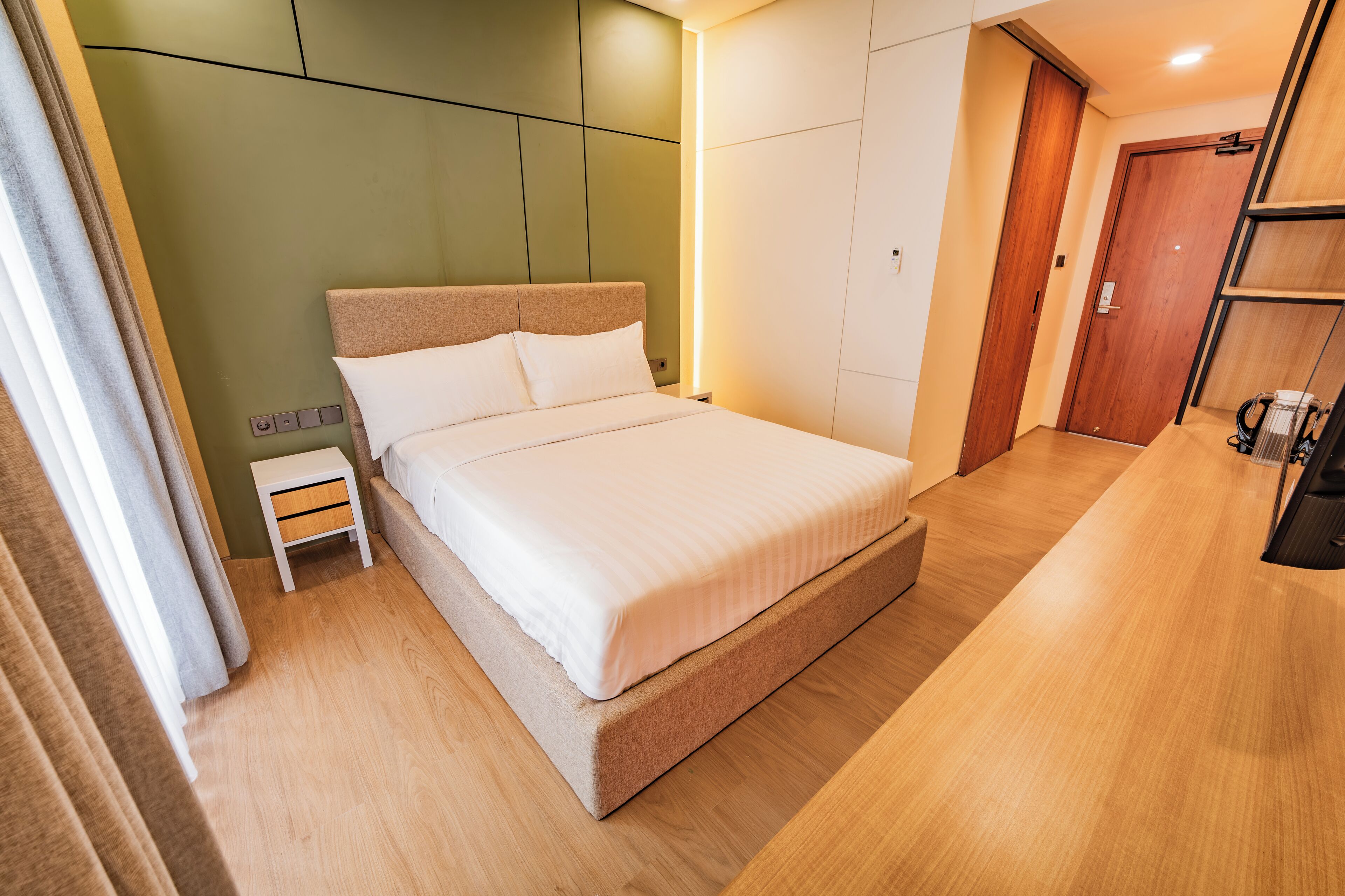 A modern hotel room with a large bed, a small bedside table, a wooden floor, and a green accent wall.