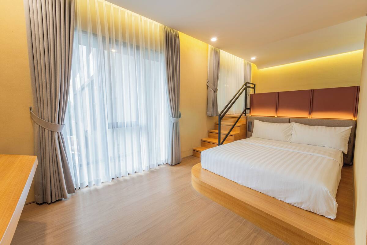 A modern bedroom with a large bed, a wooden headboard, a staircase, and sheer curtains.