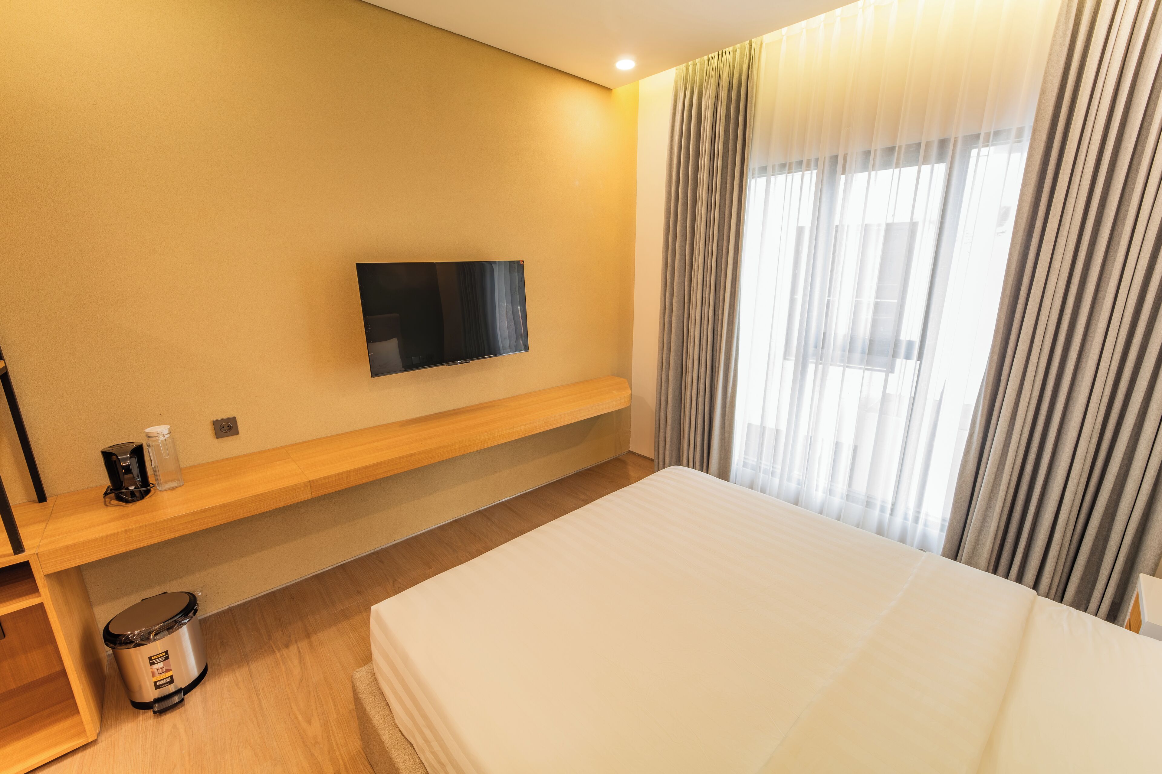 A hotel room with a bed, a television mounted on the wall, a small table with a coffee maker, and a trash bin.