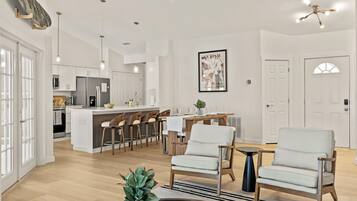 A modern kitchen with white cabinets, a central island, and pendant lighting. There is a dining area with a table and chairs, and a living room with two armchairs and a coffee table.