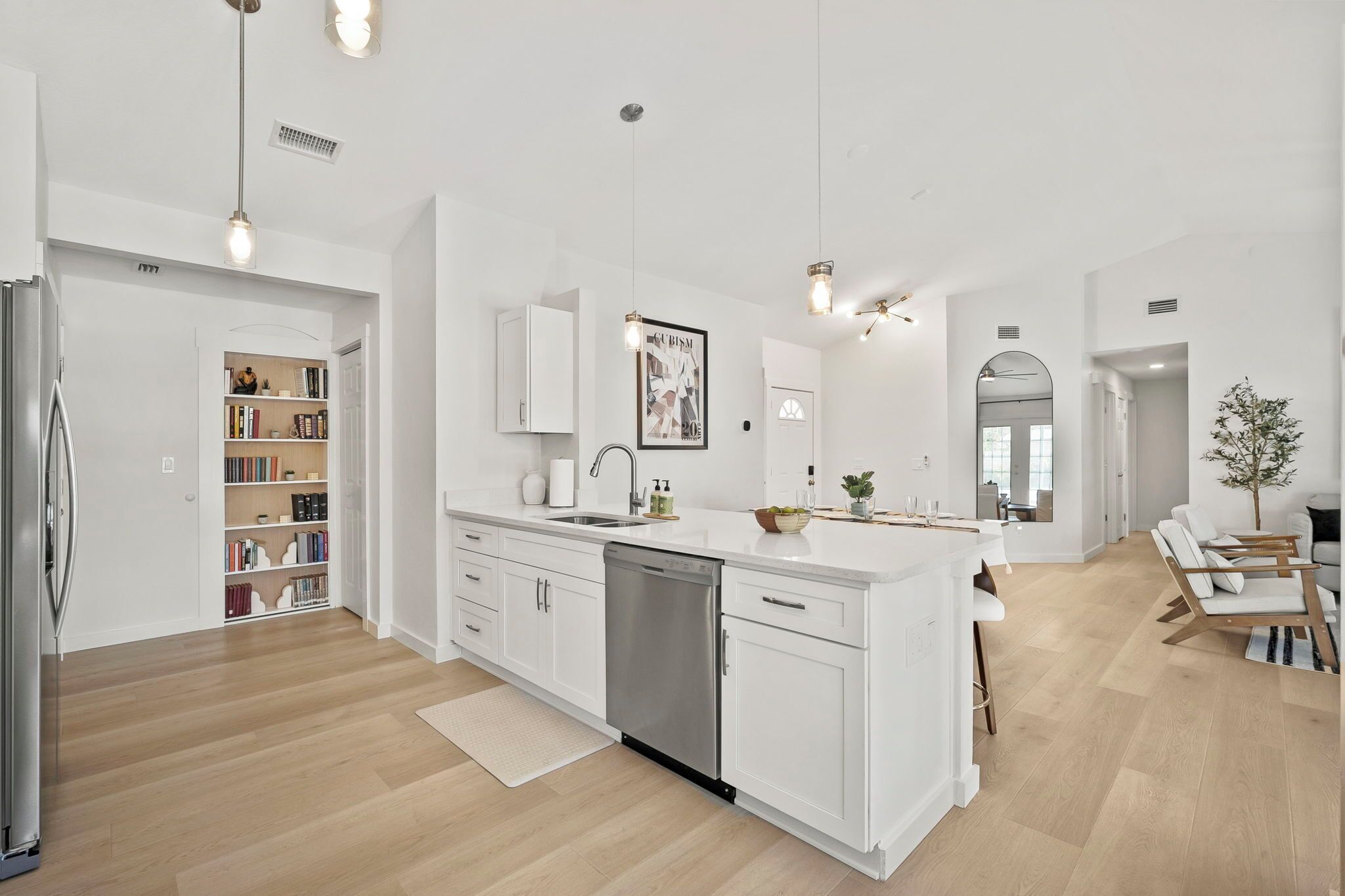 A modern kitchen with white cabinetry, a dishwasher, and a central island. There is a bookshelf with books and decorative items, and a dining area with a table and chairs.