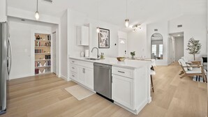 Ett modernt kök med vita skåp, diskmaskin och en köksö i mitten. Det finns en bokhylla med böcker och dekorativa föremål, samt en matplats med ett bord och stolar.