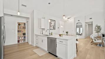 A modern kitchen with white cabinetry, a dishwasher, and a central island. There is a bookshelf with books and decorative items, and a dining area with a table and chairs.