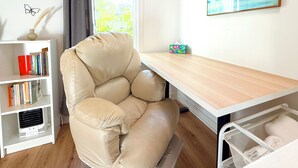 A room with a beige leather chair, a white bookshelf, a wooden desk, a framed painting, and a window with a view of trees.