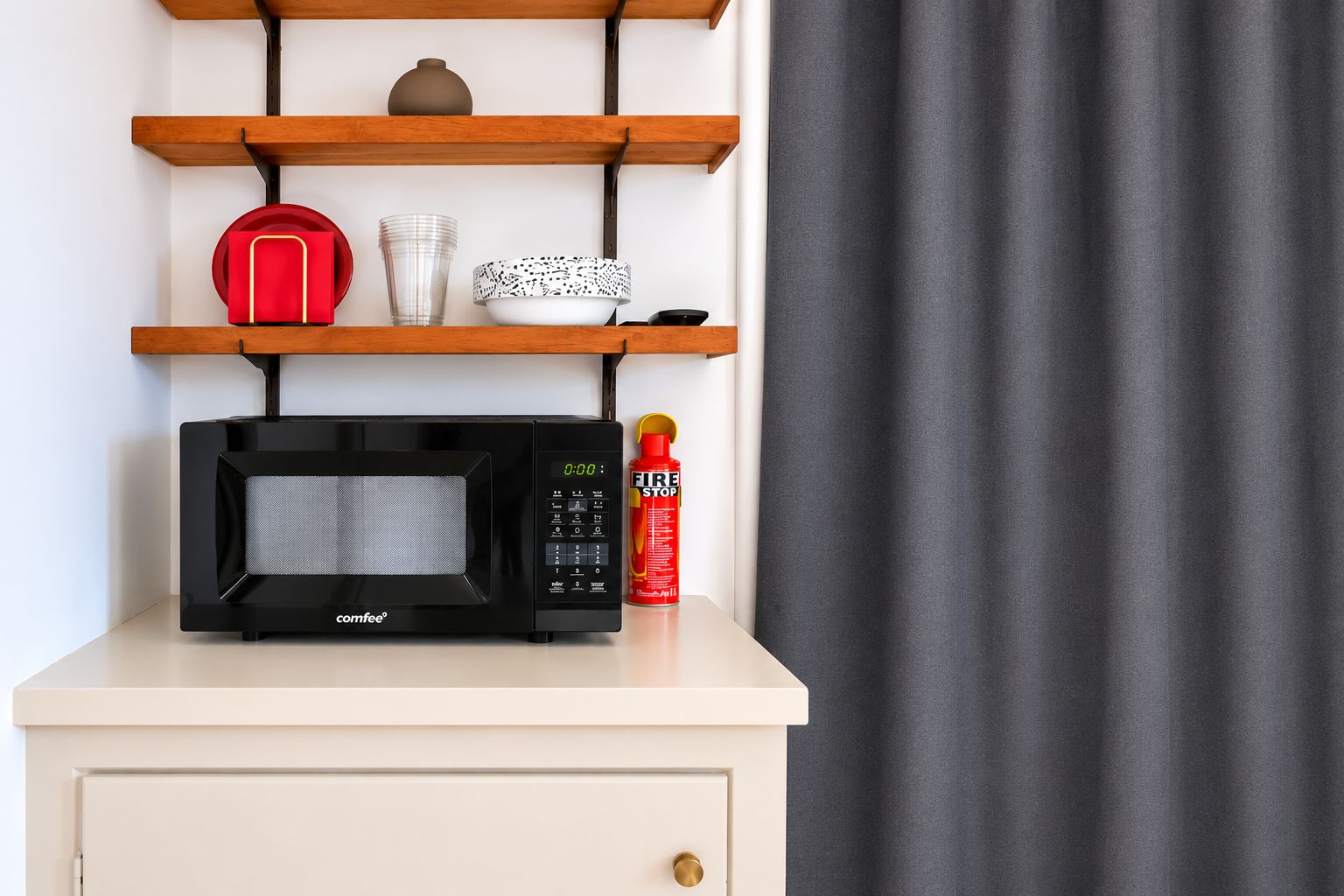 A microwave on a white cabinet with a shelf above containing a red object, a clear glass, and patterned bowls.