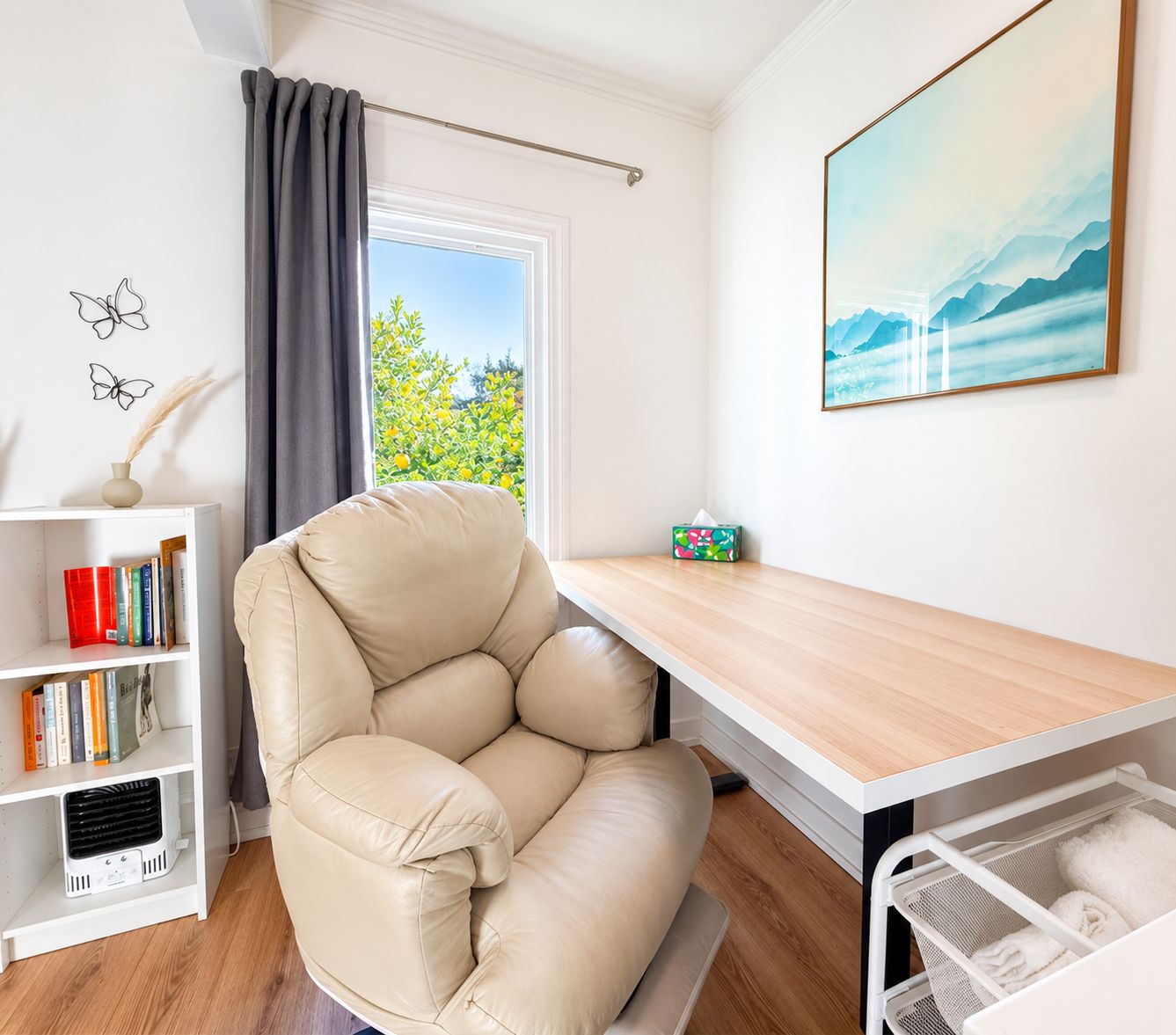 Een kamer met een beige leren fauteuil, een houten tafel, een boekenkast, een radiator en een raam met uitzicht op groen.