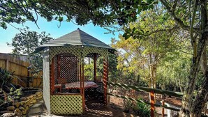 A gazebo with a lattice roof, surrounded by a wooden deck and stone pathway, set in a garden with trees and a wooden fence.