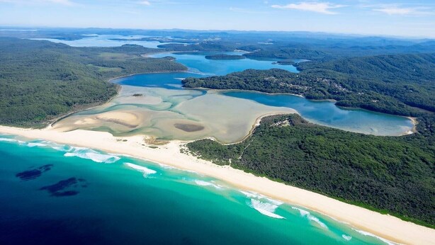 Vue aérienne d’une zone côtière avec une plage de sable, des eaux d’un bleu limpide et un paysage boisé.