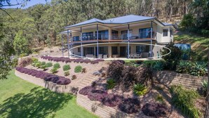Une maison moderne à deux étages, dotée d’un balcon et entourée de jardins aménagés et d’arbres.