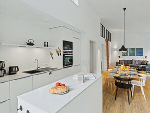 A modern kitchen with a central island, a dining area with a wooden table and chairs, and a living area with a sofa and wall art. - Haus Kiebitz (197) by Interhome (Lembruch)