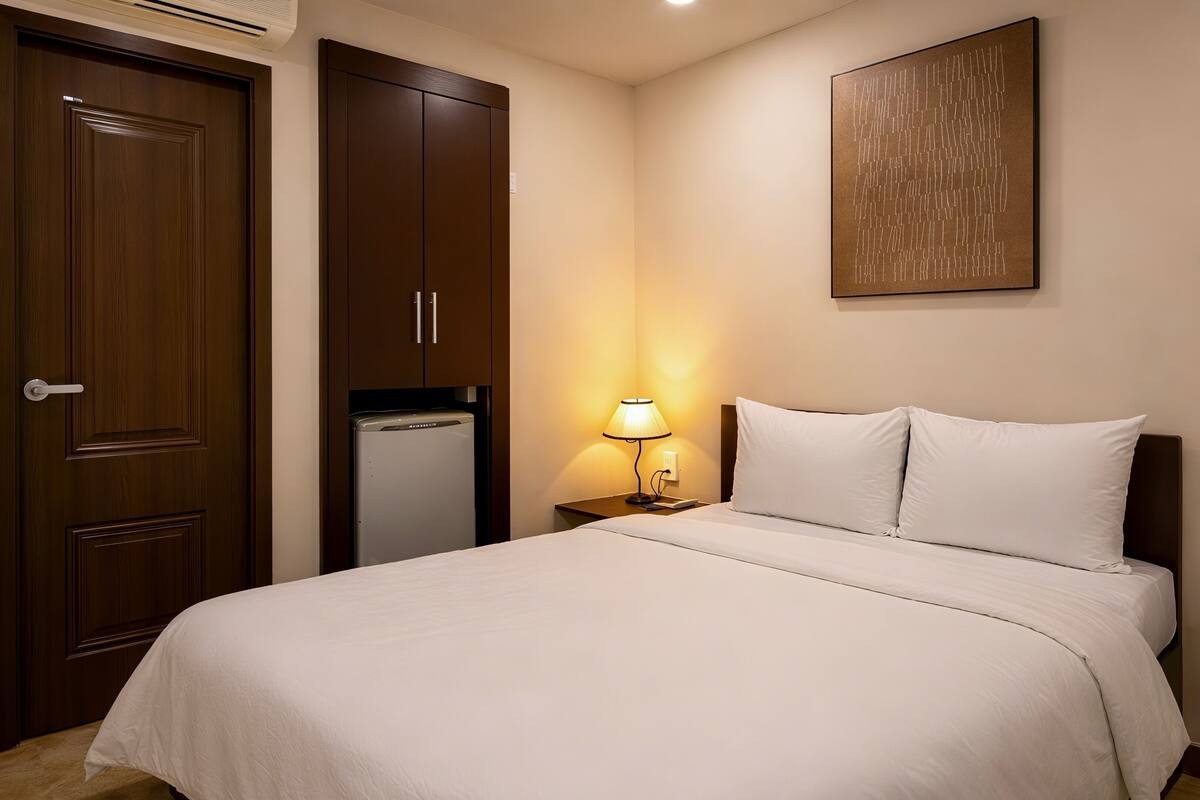 Habitació d'hotel amb un llit, una taula de nit amb una làmpada, un armari i una obra d'art penjada a la paret.