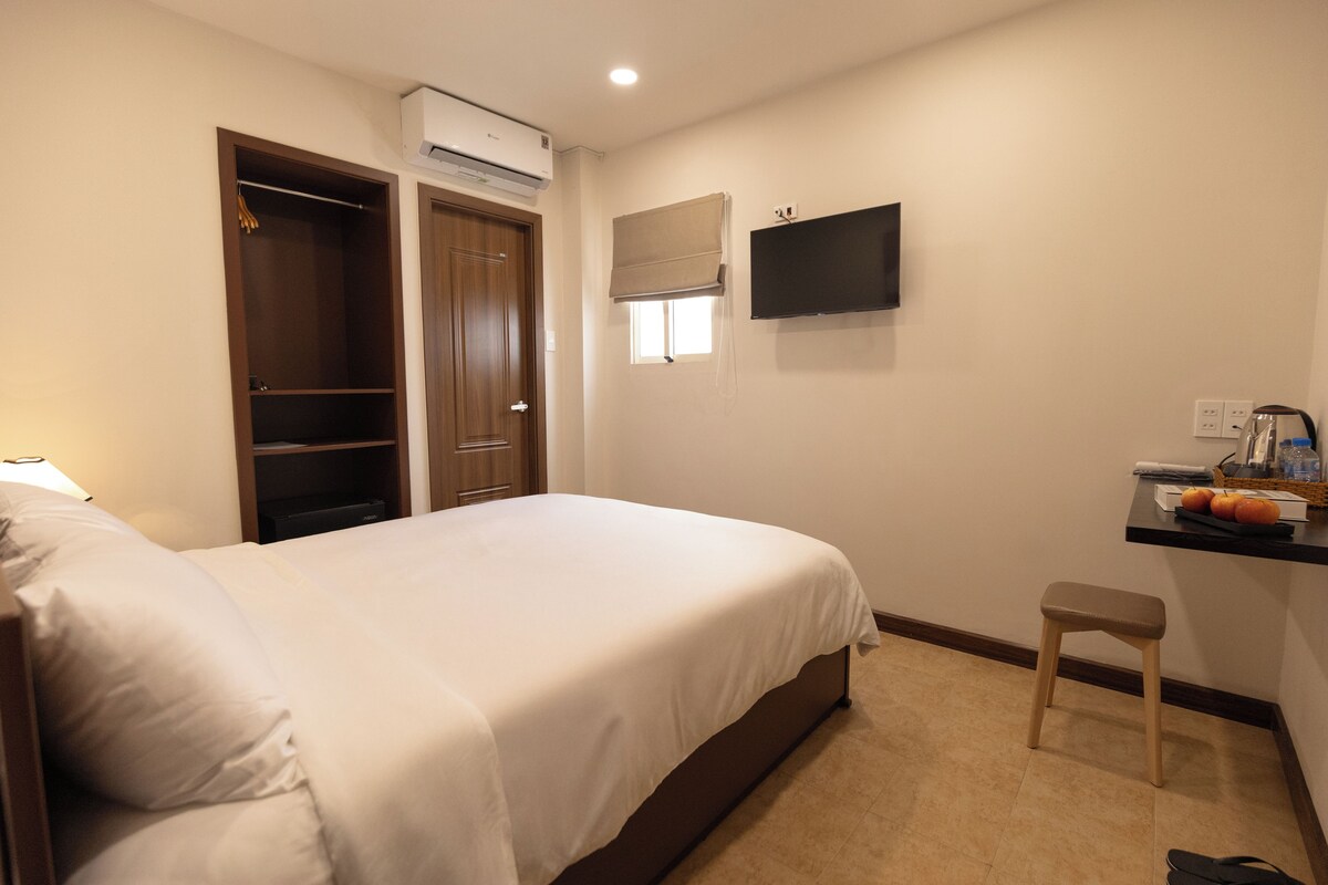 A bedroom with a bed, a wardrobe, a television, a small stool, and a desk with a basket.