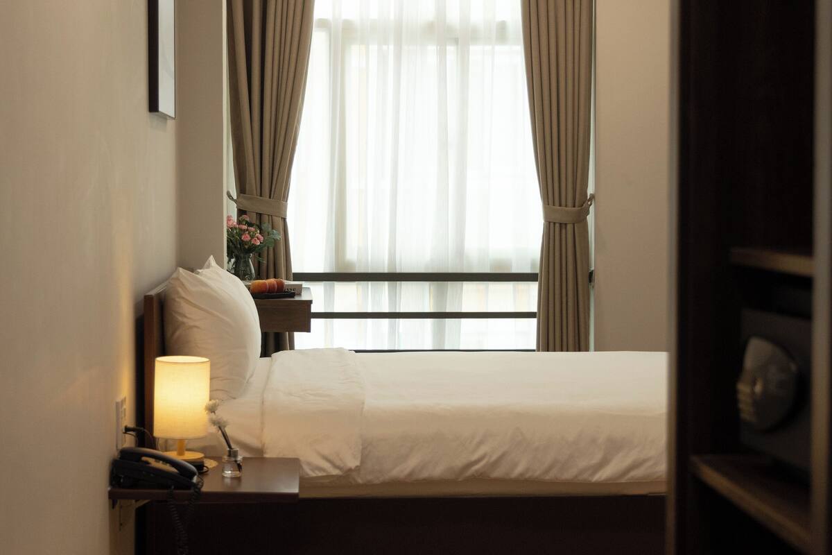 Habitació d'hotel amb llit, una tauleta de nit amb una làmpada, un telèfon i una finestra amb cortines.