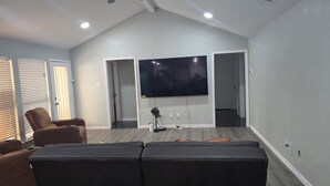 Ein Wohnzimmer mit Flachbildfernseher an der Wand, einem Ecksofa, einem Liegestuhl und einem kleinen Tisch mit Lampe.