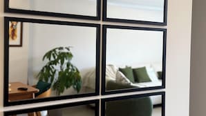 Ein an der Wand befestigter Spiegel mit schwarzem Rahmen, der einen Raum mit einer Couch, einer Zimmerpflanze und einem Holztisch reflektiert.