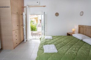 Une chambre avec une couvre-lit verte, des oreillers blancs et une table de chevet avec une lampe. On y trouve une armoire en bois et une porte-fenêtre coulissante donnant sur un balcon.