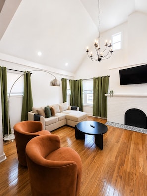 A modern living room with a sectional sofa, two armchairs, a coffee table, a fireplace, a TV mounted on the wall, and a chandelier. - Dogs Welcome!  Fun, Stylish Detroit Tudor w/ Fenced Yard! (Detroit)