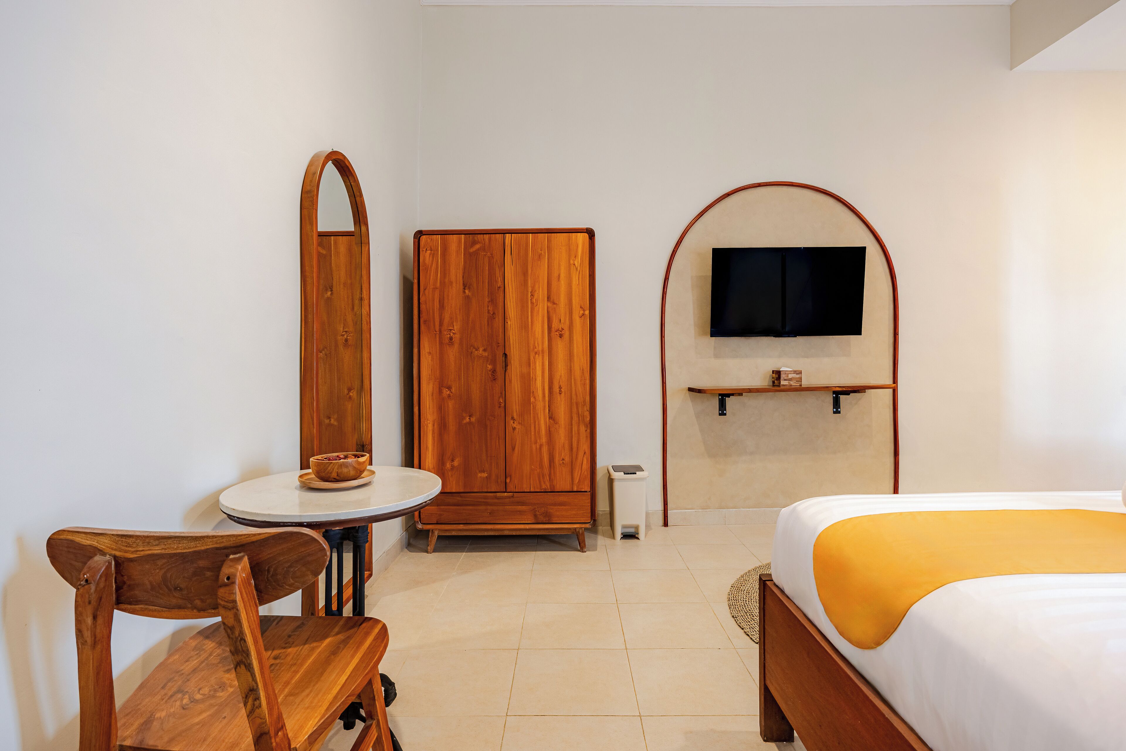 A hotel room with a wooden wardrobe, a round table with a bowl, a bed with a yellow blanket, and a mounted TV.