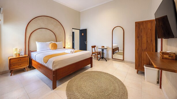 A modern hotel room with a wooden bed, a small round rug, a desk with a mirror, and a TV. - Cove Chara (Jimbaran)