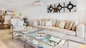 Uma sala de estar moderna com uma mesa de centro de vidro, sofá bege e uma área de jantar com mesa e cadeiras de madeira.