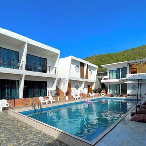 Un hotel moderno con piscina, varios balcones y vista a una colina.