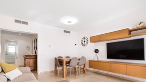 Een moderne woonkamer met een bank, eettafel en stoelen. Een televisie hangt aan de muur en erboven hangt een klok.