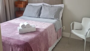 Una cama bien hecha con una manta rosa, una mesita de noche de madera con espejo y una silla blanca en una habitación moderna con ventilador de techo.