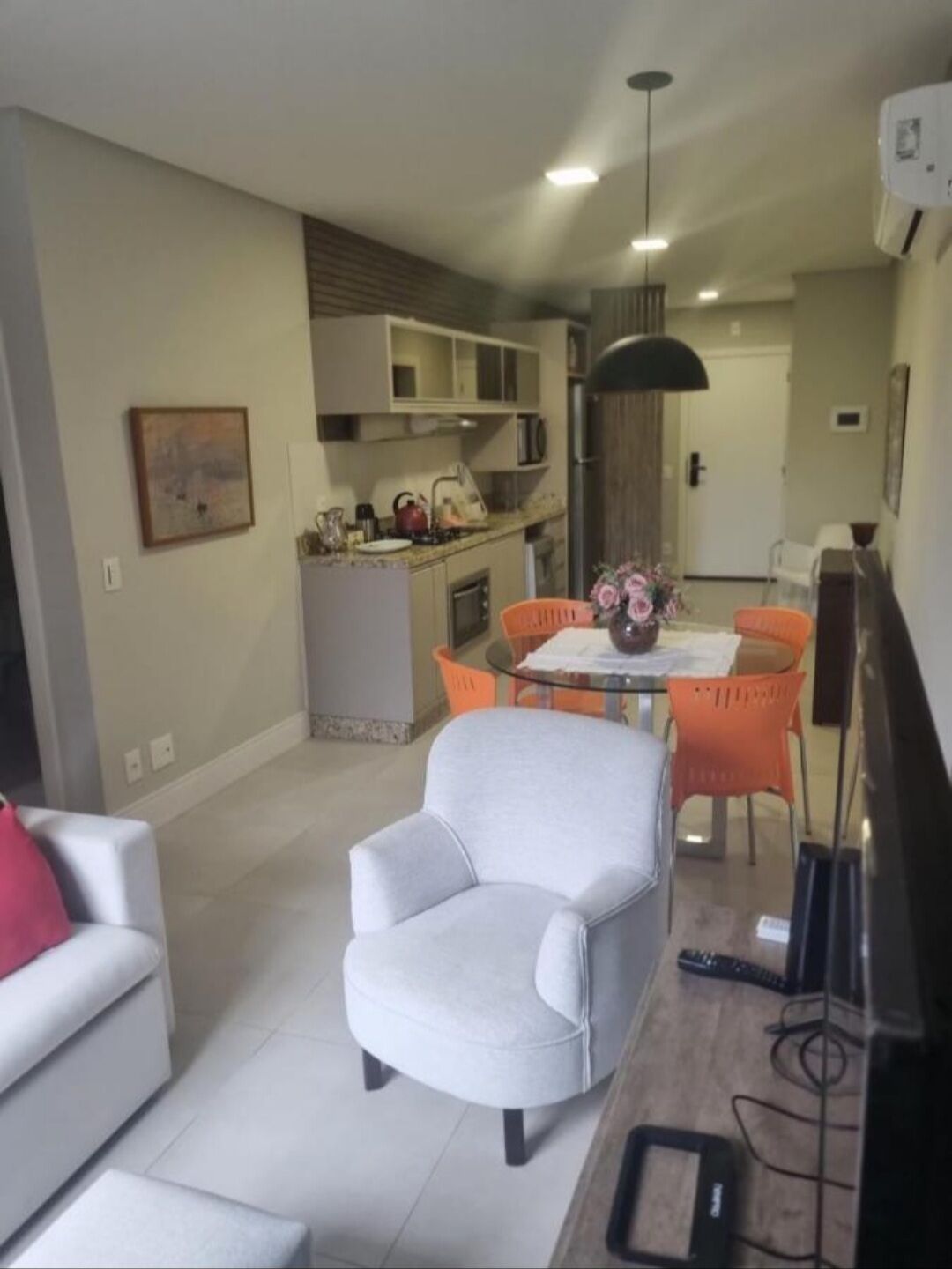 Una sala moderna con un sillón gris, un sofá blanco y una mesa de comedor con sillas naranjas. Se ve una cocina al fondo con un microondas y una tetera.