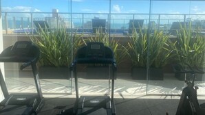 Un gimnasio con cintas de correr y plantas en macetas, amplias ventanas con vista a la ciudad.