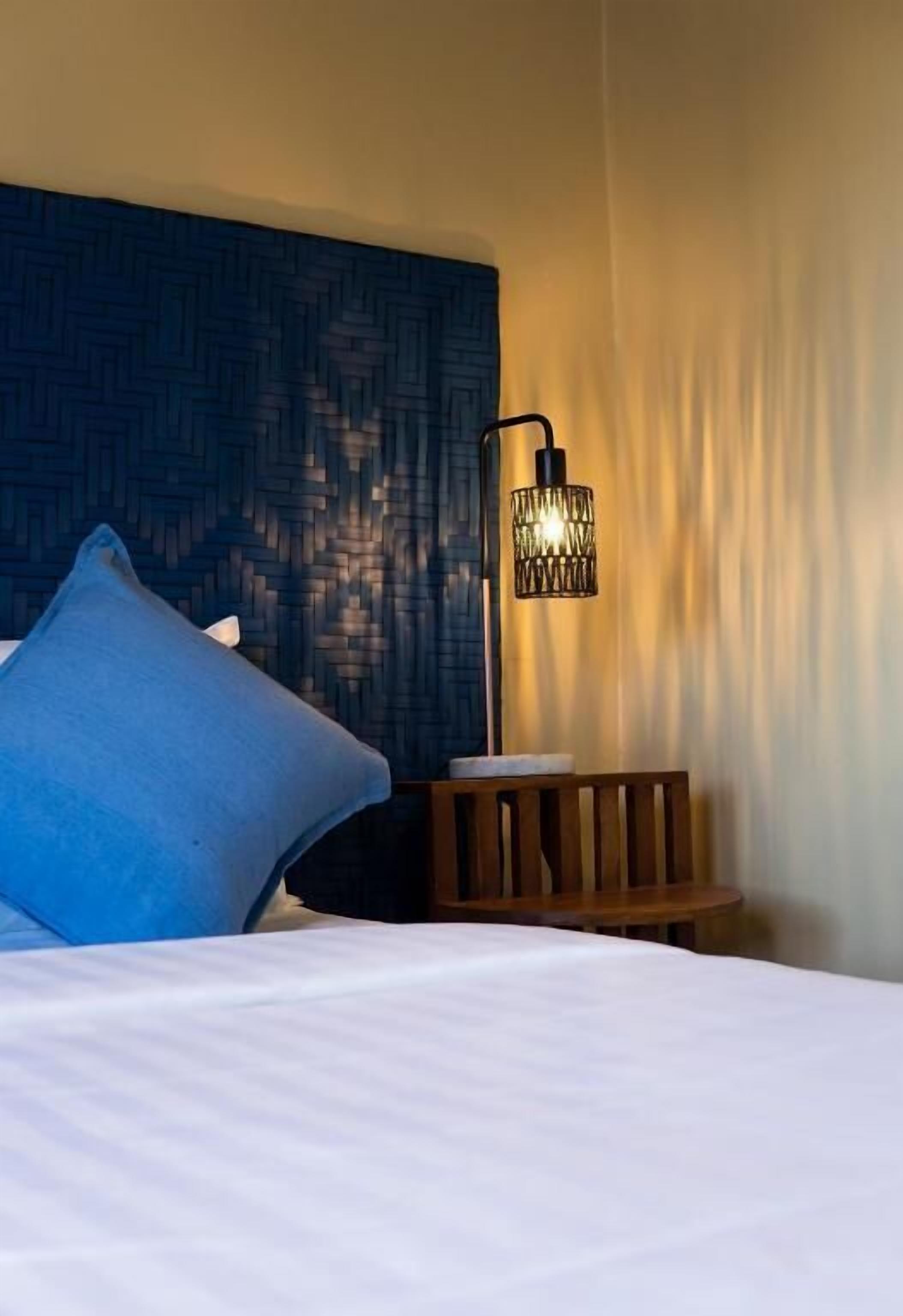 A neatly made bed with a blue pillow, a wooden nightstand, and a wall-mounted lamp.