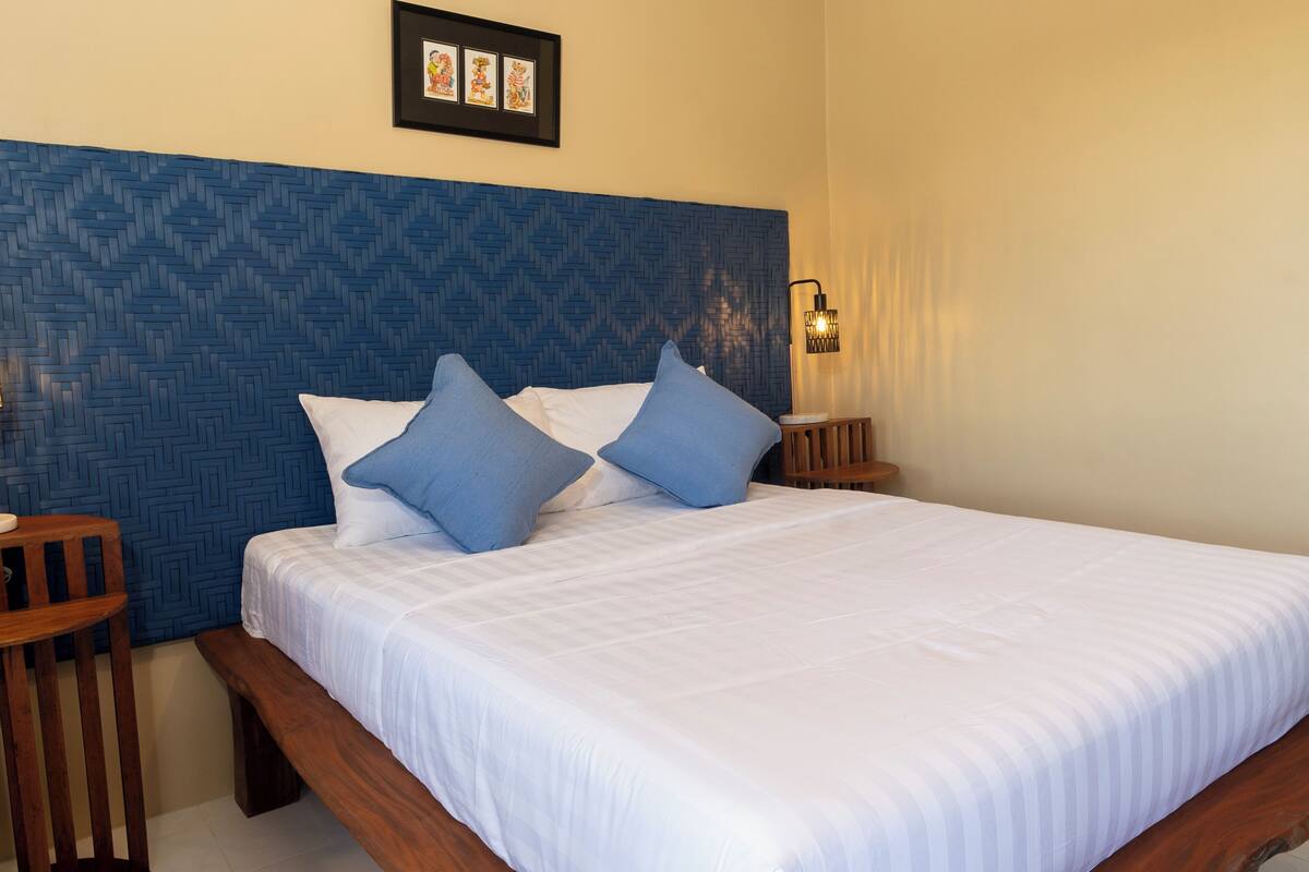 A neatly made bed with white bedding and blue pillows, a wooden headboard with a blue geometric pattern, and a wall-mounted light fixture.