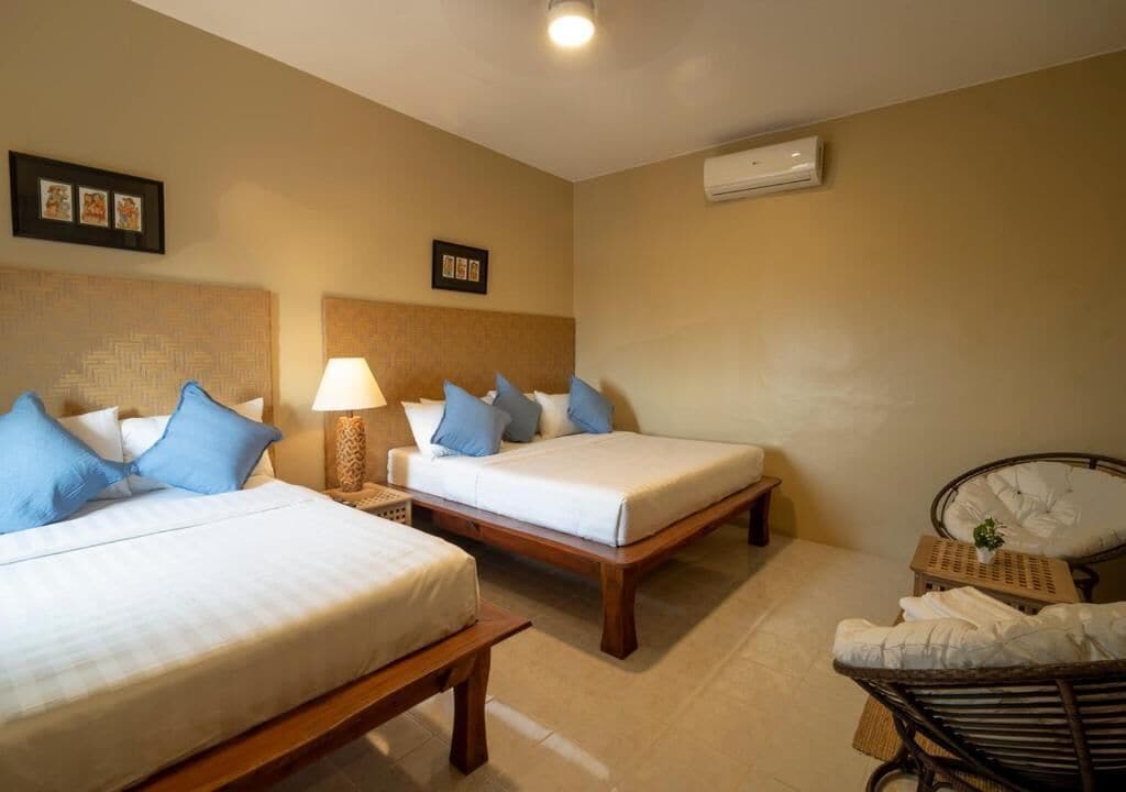 A hotel room with two single beds, a bedside table with a lamp, a chair with a cushion, and a small table with a plant.