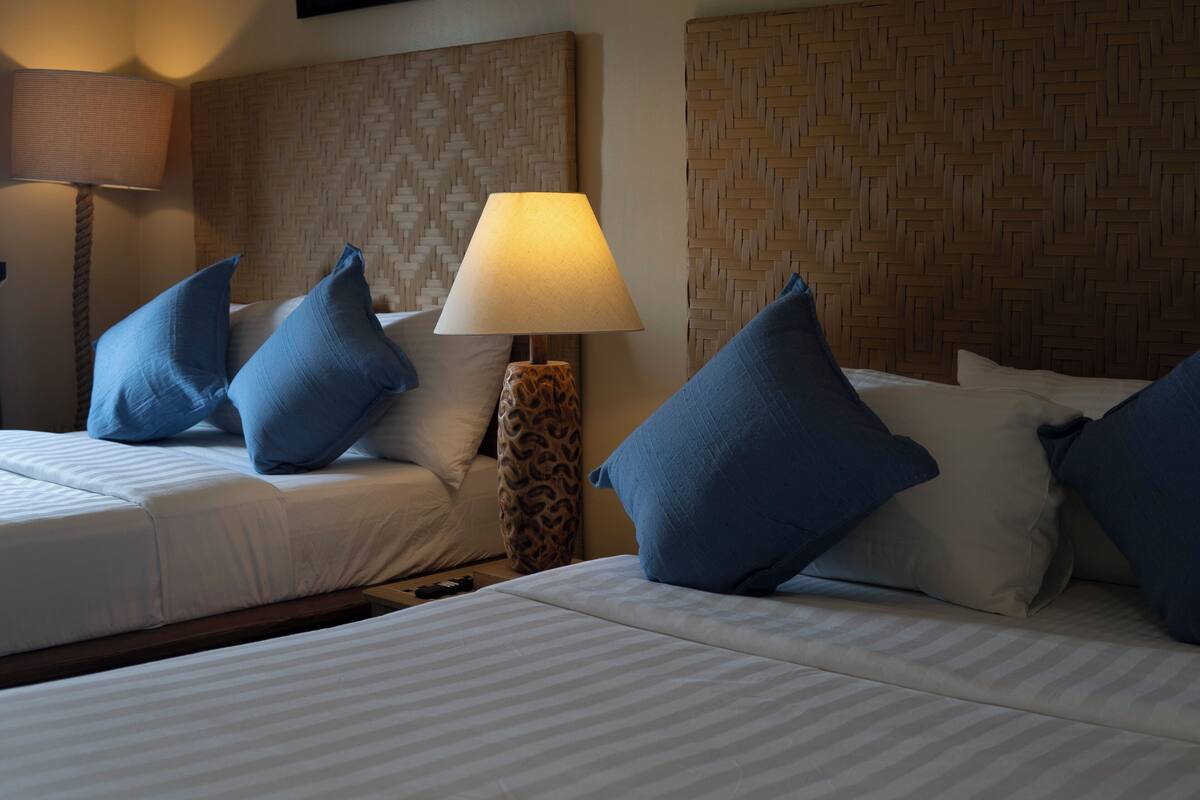 A hotel room with two beds, a woven headboard, a lamp, and blue pillows.