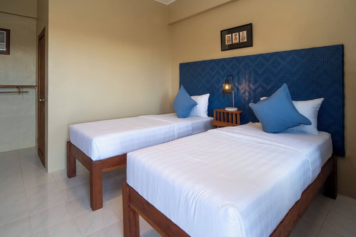 A hotel room with two single beds, a nightstand with a lamp, and a door leading to another room.