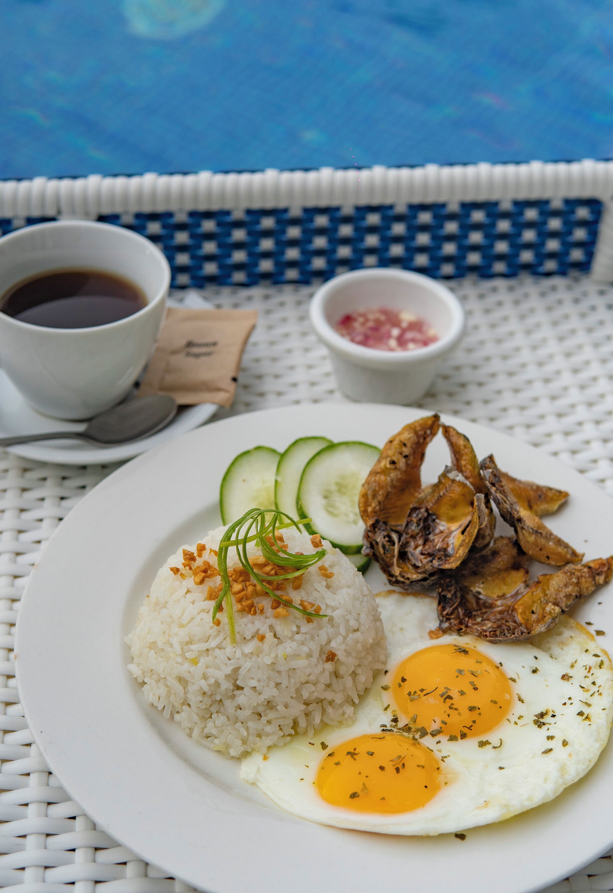 A plate of food with rice, fried egg, and fried plantains, accompanied by a cup of coffee and a small bowl of sauce, on a white table with a blue cushion.