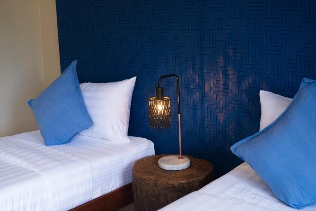 Due letti con lenzuola bianche e cuscini blu, un comodino in legno tondo con una lampada da terra e una parete azzurra con texture.